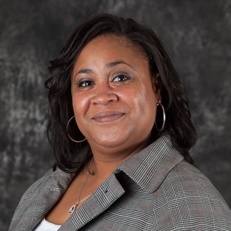 Professional headshot of a woman in a gray blazer on a dark background